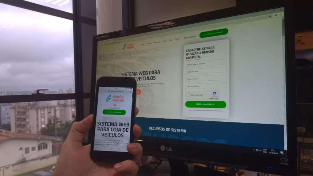 Mão segurando celular com o sistema web para loja de veículos, diante de um monitor com o site aberto
