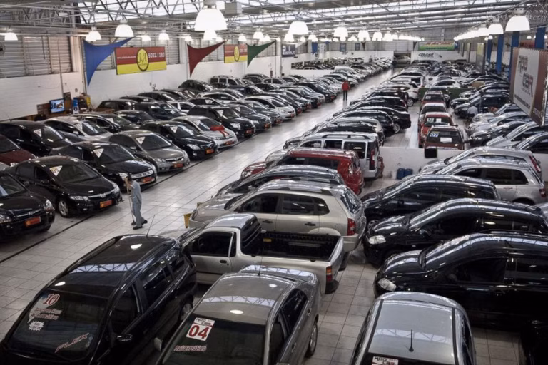 Carros estacionados em fileiras dentro de um pátio coberto, com bandeiras penduradas no teto