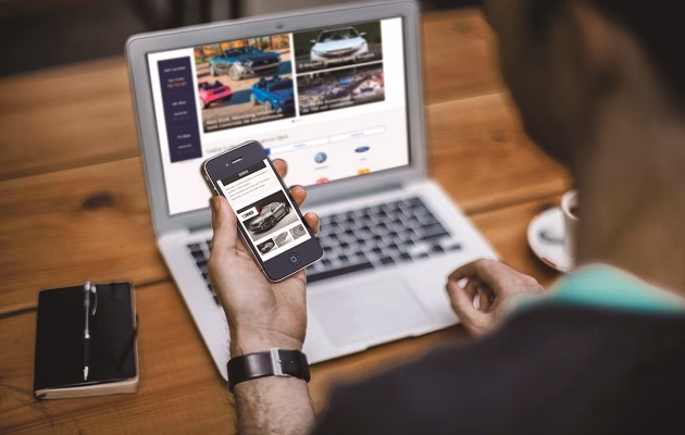 Pessoa olhando anúncios de carros no celular, com notebook aberto em site de veículos na mesa