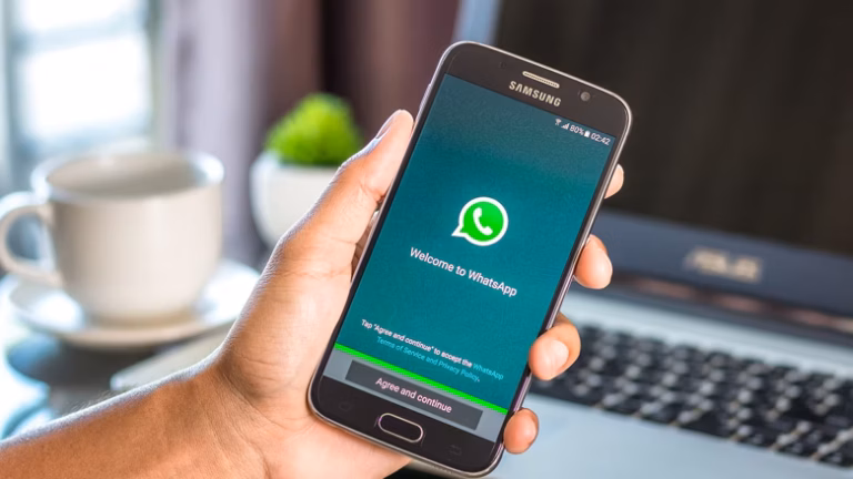Mão segurando celular com a tela de boas vindas do WhatsApp, notebook e xícara ao fundo