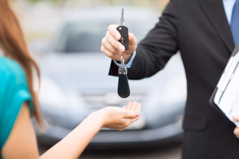 Homem de terno entregando a chave de um carro para uma mulher, com carro prata ao fundo