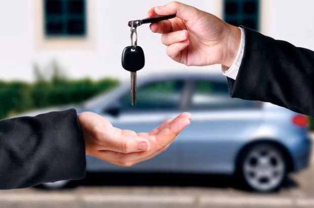 Mão de terno entregando a chave de um carro a outra pessoa, com sedã azul ao fundo