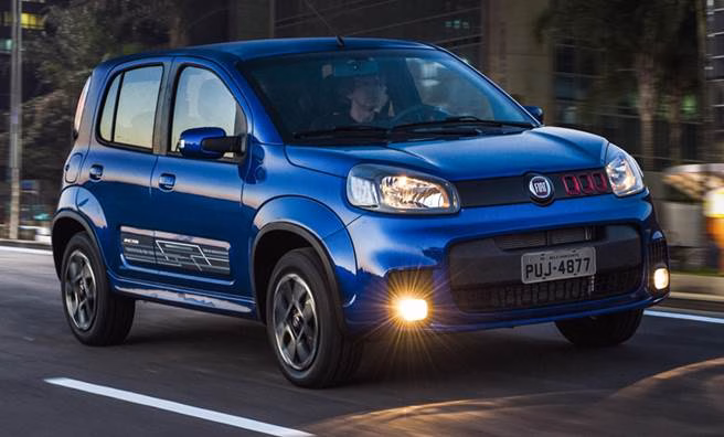 Fiat Uno azul com faróis acesos rodando em rua ao entardecer