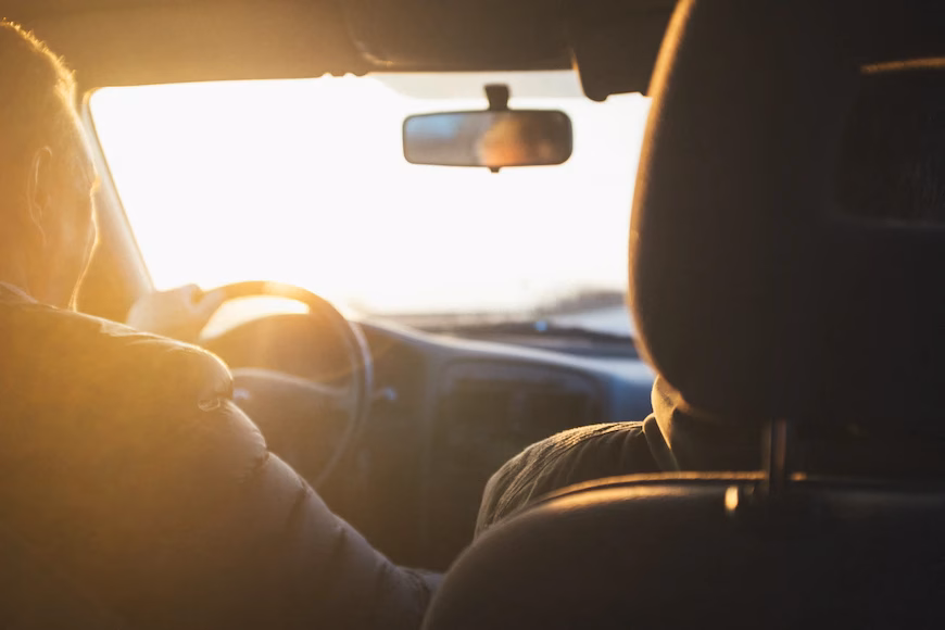 Motorista e passageiro vistos do banco de trás, com sol entrando pelo para-brisa