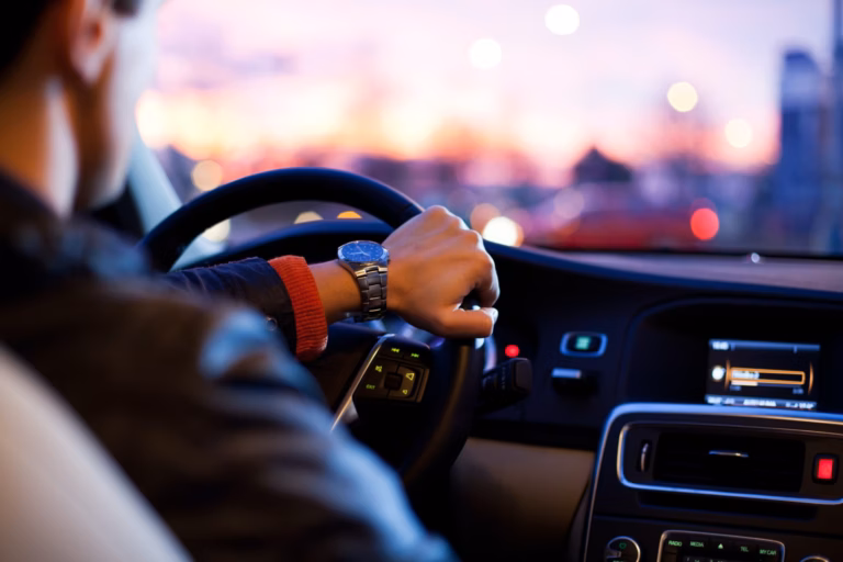 Motorista com relógio no pulso dirigindo, luzes da cidade desfocadas ao fundo