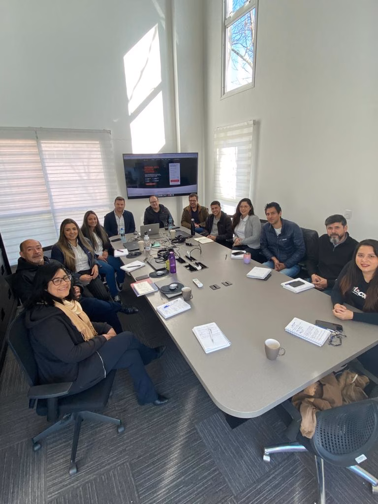 Grupo de pessoas reunidas ao redor de uma mesa de reunião durante treinamento, com tela ao fundo