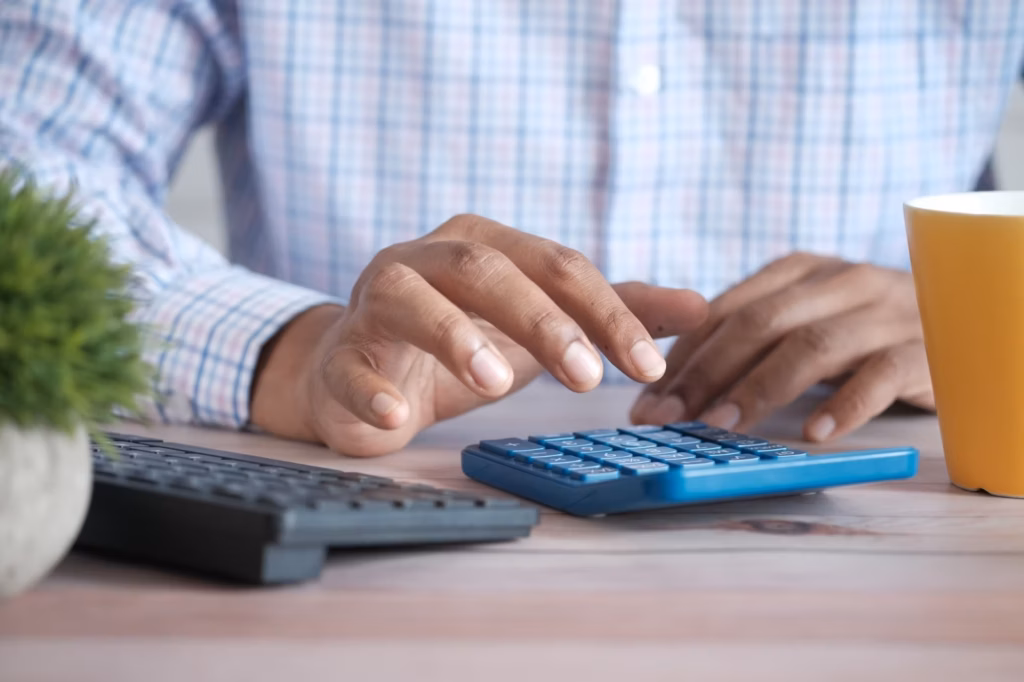 Homem de camisa xadrez fazendo contas em uma calculadora azul ao lado do teclado