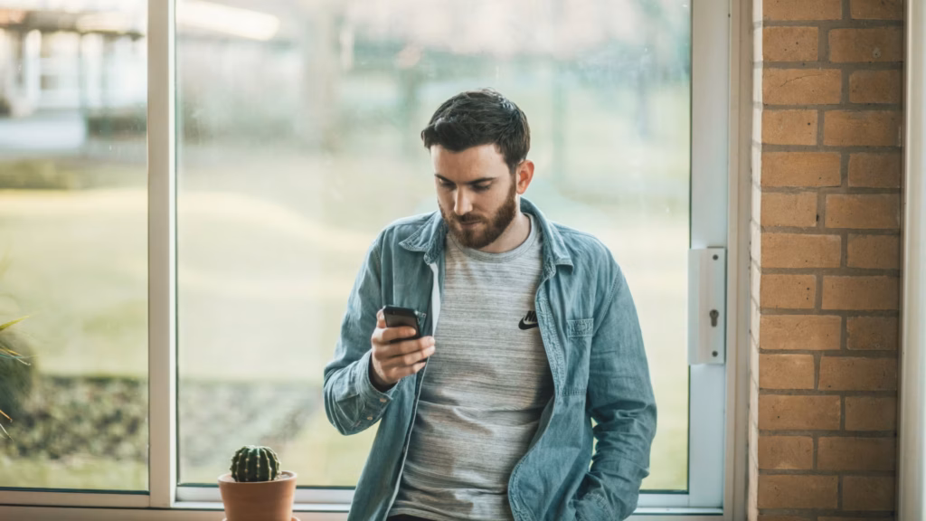 Homem de barba e jaqueta jeans olhando o celular perto de uma janela