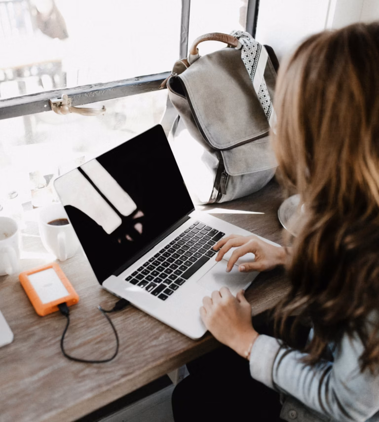Mulher usando notebook em uma mesa com mochila e HD externo ao lado
