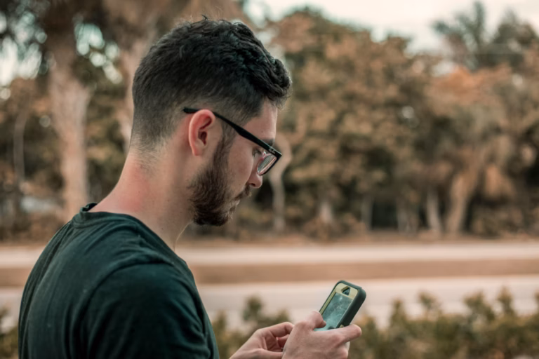 Homem de barba e óculos olhando o celular ao ar livre, com árvores ao fundo