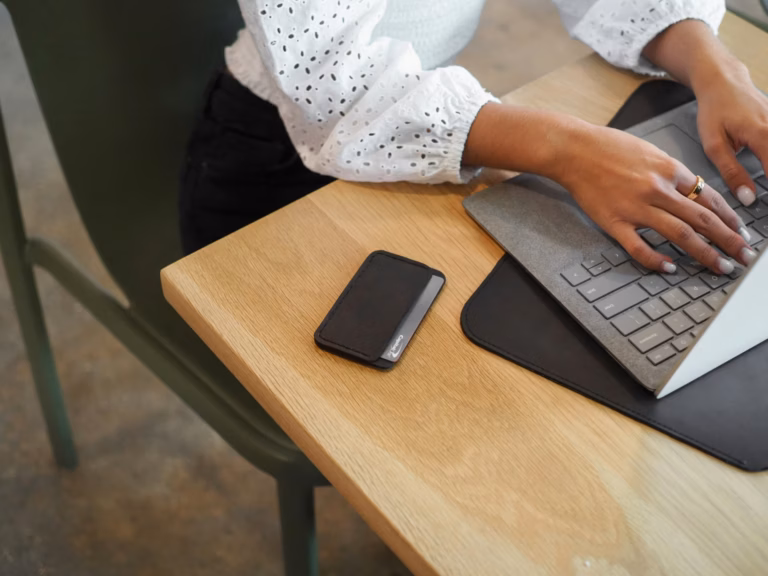 Mãos digitando em um teclado sem fio sobre mesa de madeira, com celular ao lado