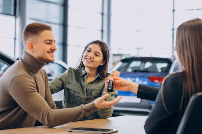 Casal sorridente recebendo a chave do carro de uma vendedora em mesa de showroom
