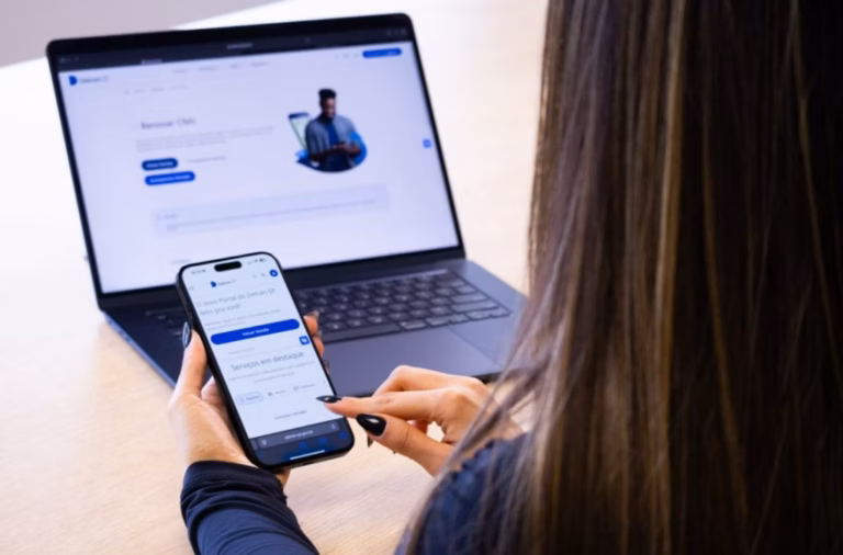Mulher usando o celular diante de um notebook aberto em uma página com foto de pessoa