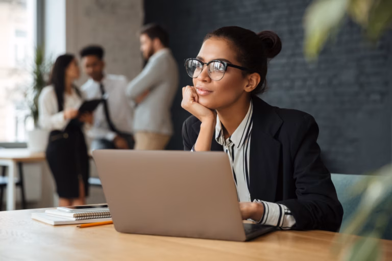 Mulher de óculos pensativa diante de um notebook, com colegas conversando ao fundo