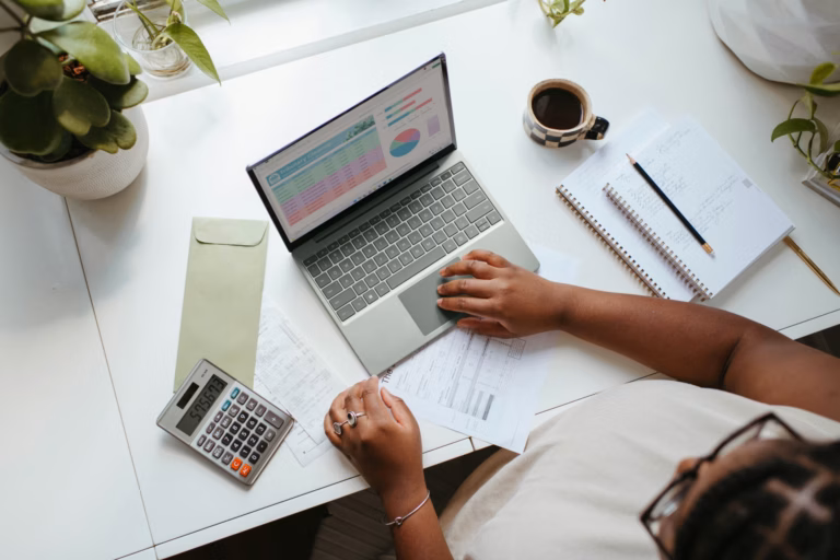 Pessoa analisando gráficos no notebook, vista de cima, com calculadora, caderno e café na mesa