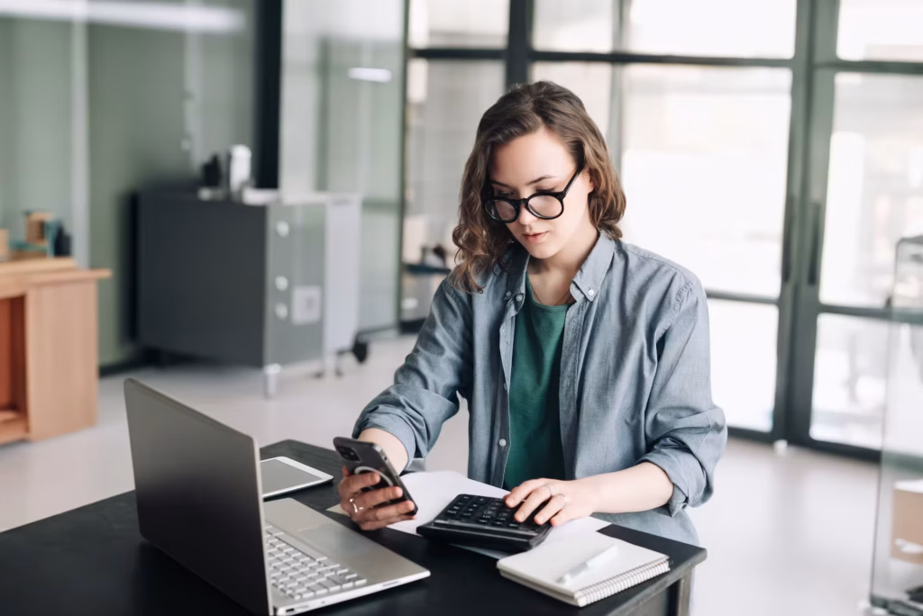 Mulher de óculos fazendo contas na calculadora com celular e notebook na mesa