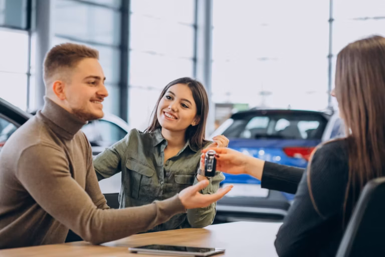 Casal sorridente recebendo a chave do carro de uma vendedora, com carro azul ao fundo