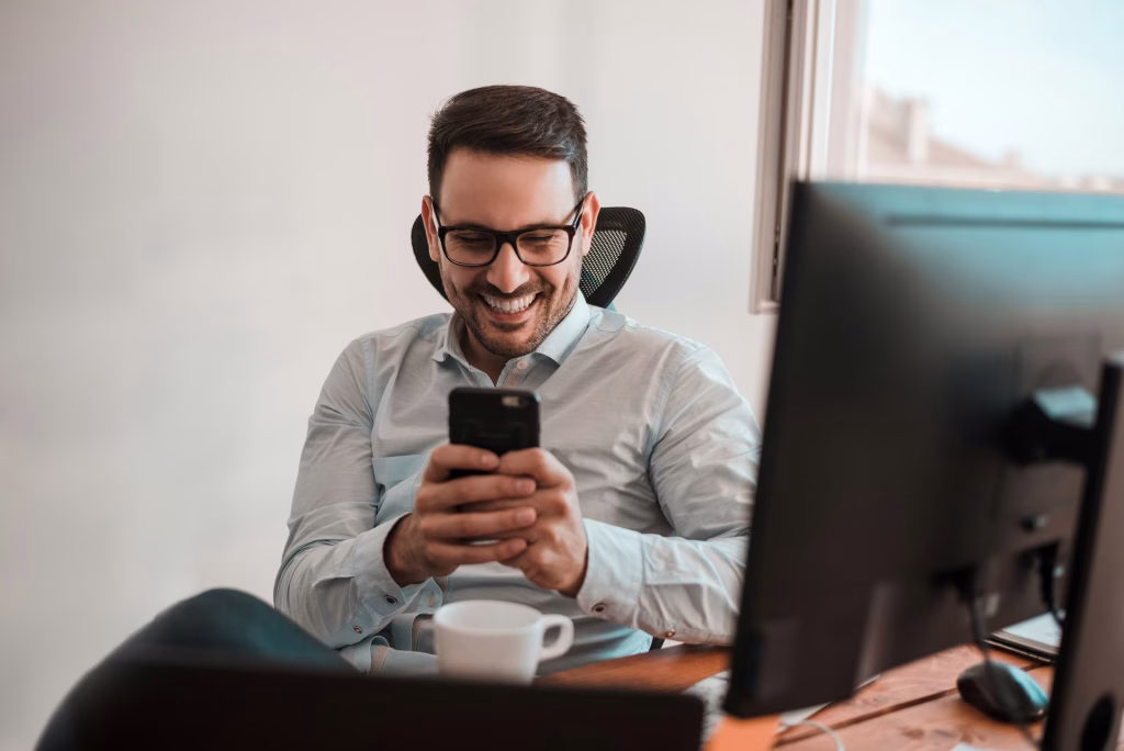 Homem de óculos sorrindo enquanto usa o celular no escritório