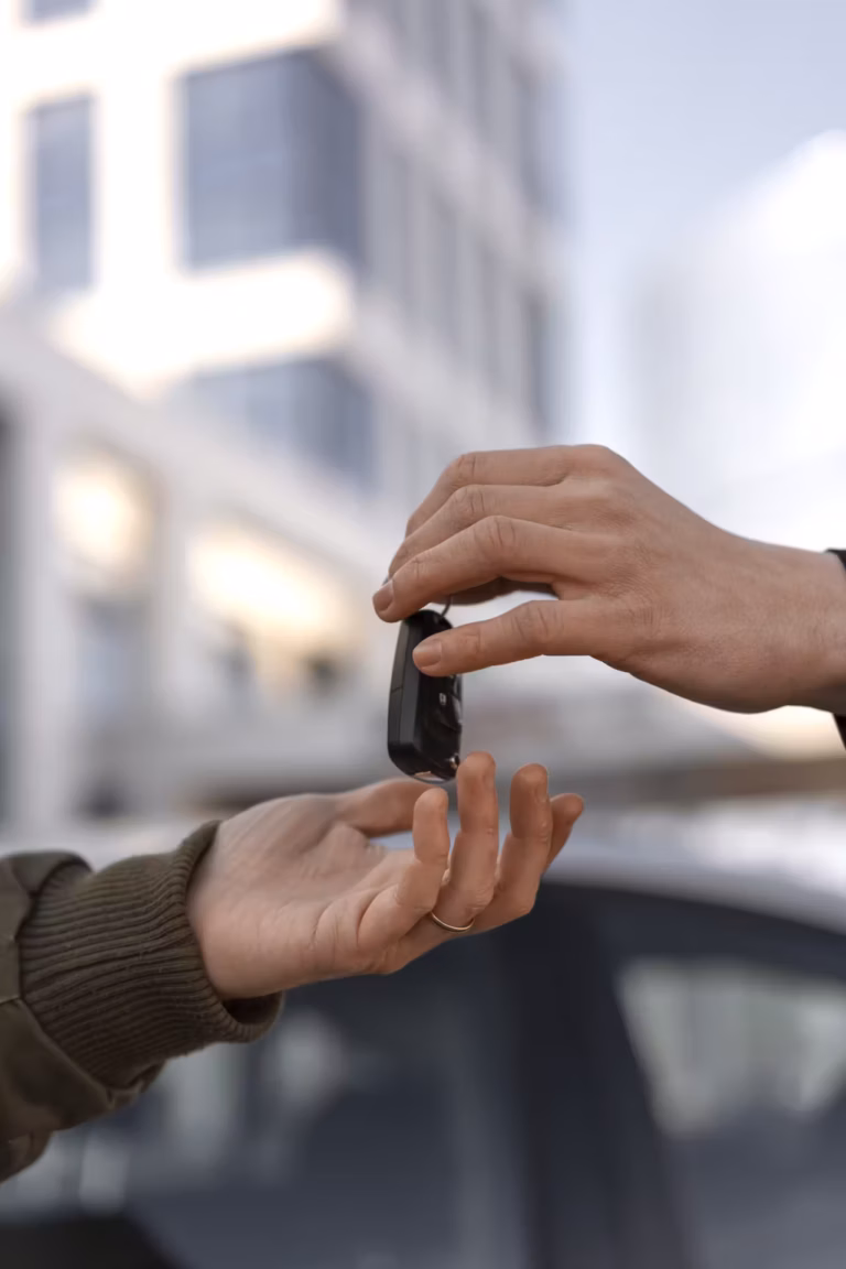 Mão entregando a chave de um carro para outra mão, com prédio desfocado ao fundo