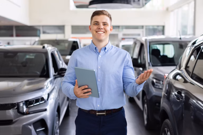 Vendedor sorridente de camisa azul segurando um tablet entre carros no showroom da revenda